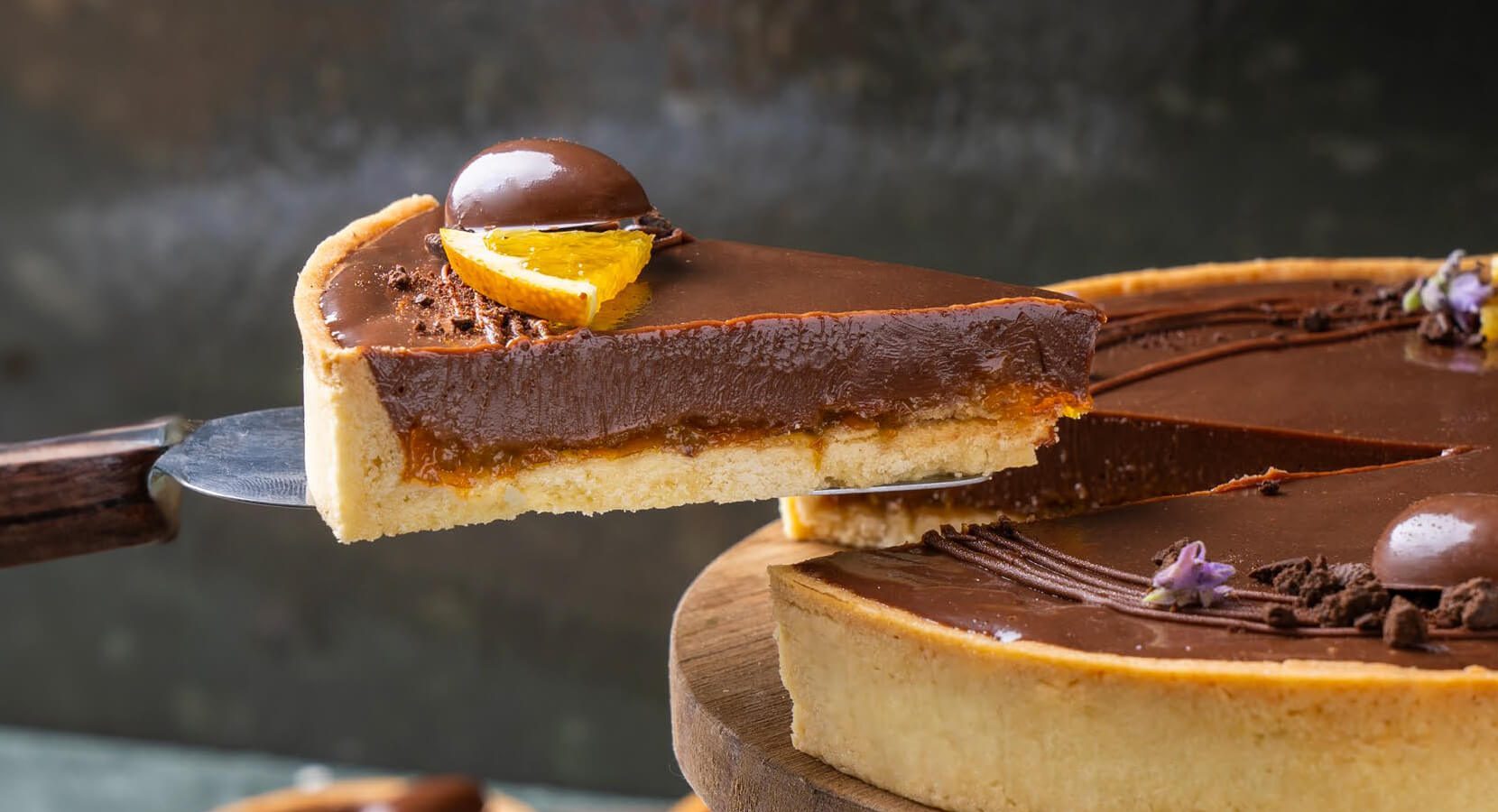 A imagem mostra uma tartelete de chocolate ao leite sendo cortada e servida. A tartelete tem uma base de massa crocante, com recheio de caramelo e uma camada superior de ganache de chocolate ao leite, brilhante e lisa. A decoração inclui pedaços de laranja cristalizada, uma esfera de chocolate e pequenos detalhes de flores comestíveis roxas e pedaços de chocolate ralado. O fundo é desfocado, destacando o doce no centro da composição. A tartelete está posicionada sobre uma base de madeira rústica, realçando sua aparência artesanal e apetitosa.