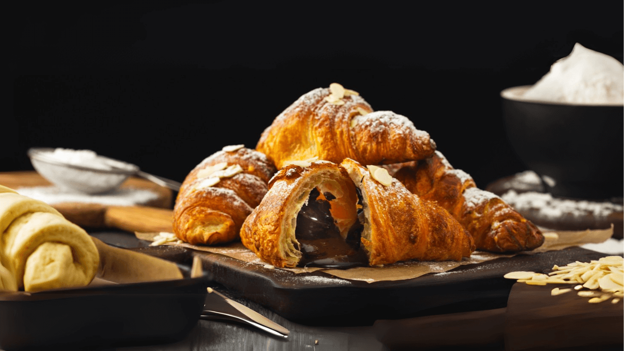 Imagem de croissants dourados e bem folhados dispostos sobre uma tábua de madeira escura, com um croissant ao centro cortado ao meio, revelando um recheio cremoso de chocolate que escorre delicadamente. O topo dos croissants é polvilhado com açúcar de confeiteiro e decorado com lascas de amêndoas, criando um visual apetitoso. Ao fundo, há elementos de panificação, como farinha e utensílios, sugerindo um ambiente de preparo artesanal.